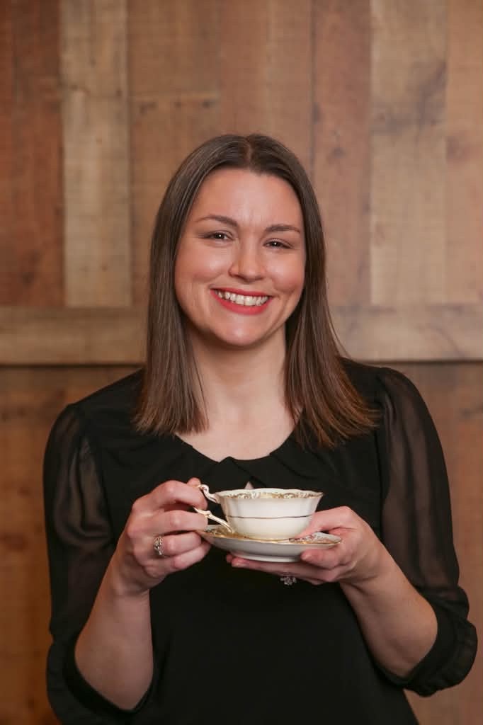 Photo of Nicole Wilson smiling and holding a gold-rimmed teacup and saucer in front of a weathered wood wall